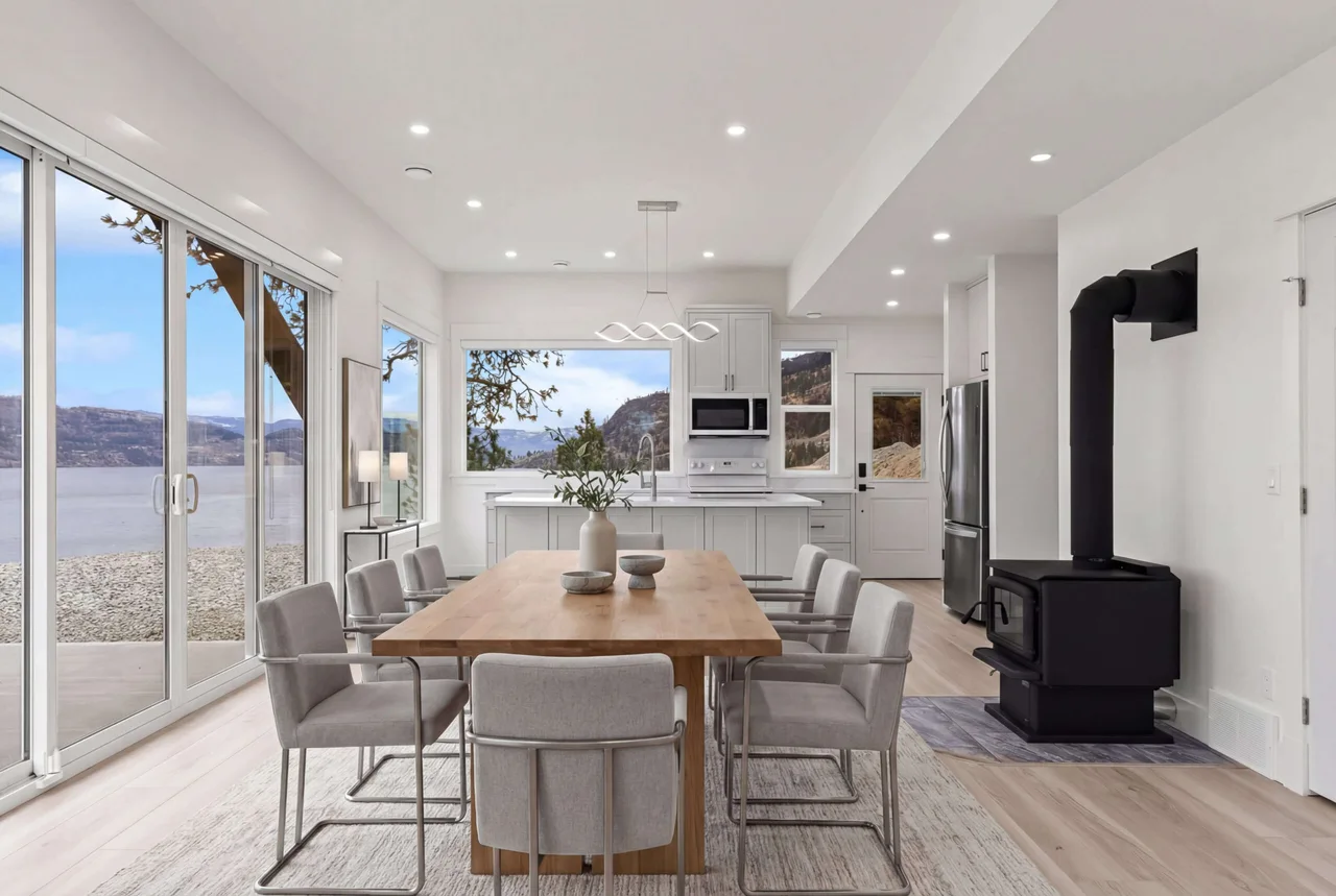 An open kitchen and dining area with a long wood table, a black wood stove and sliding doors onto the lake