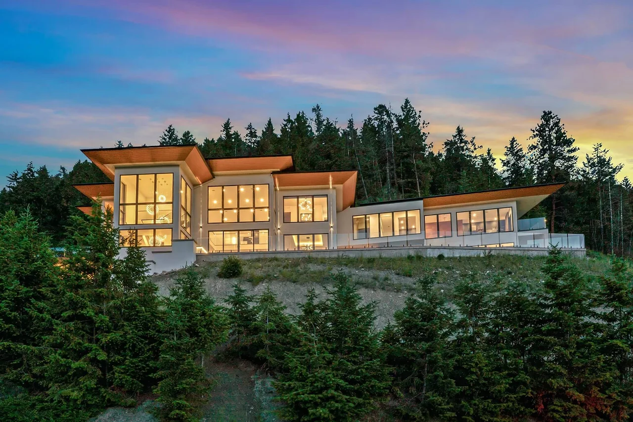 A large modern home with warm-lit walls of glass on a forested hillside at sunset