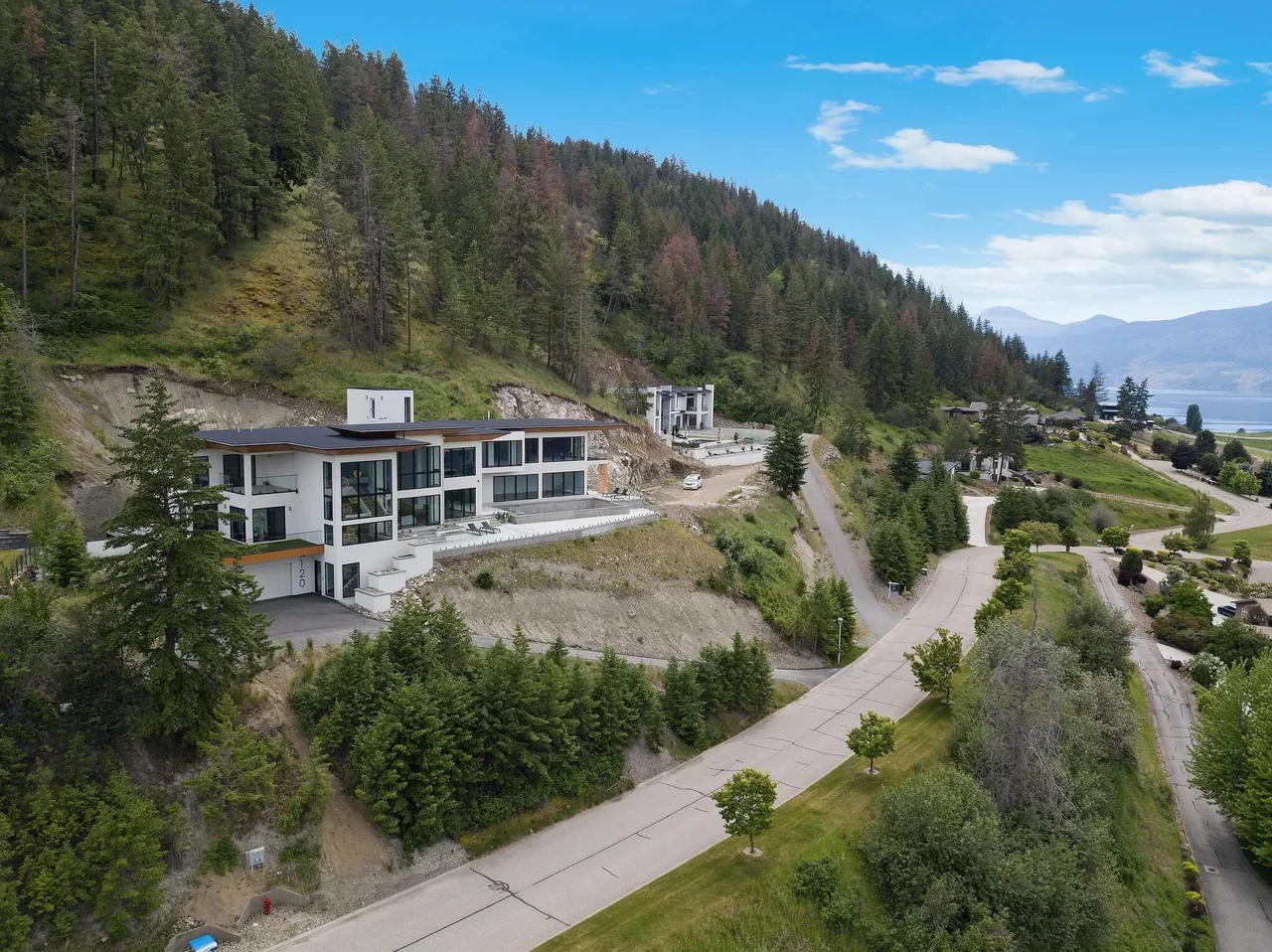 An aerial view of a modern home on a hillside lot above a winding road, with the lake in the distance