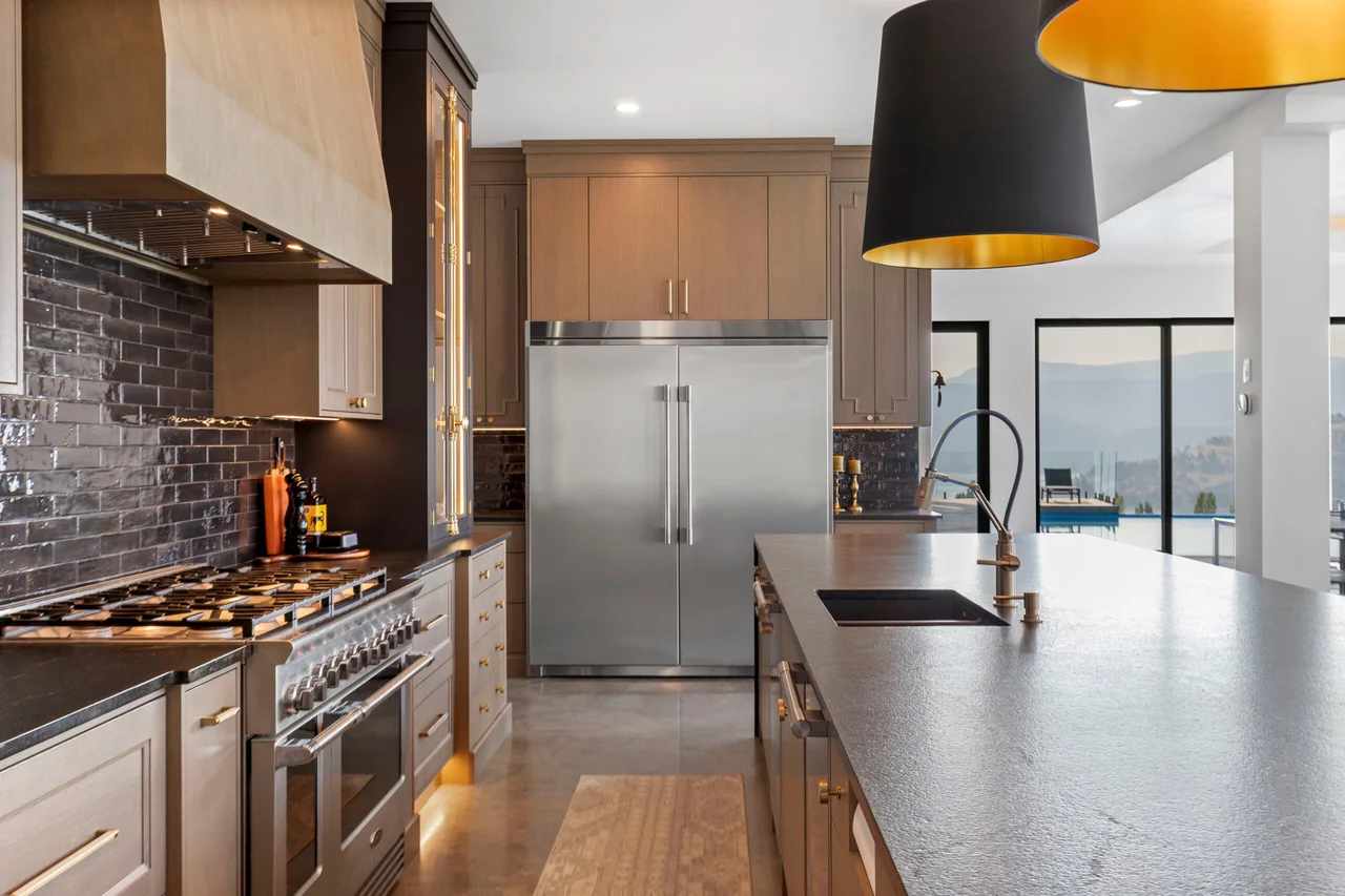 A kitchen with a dark tile backsplash, a stainless range and fridge, a stone island and large black and gold pendants