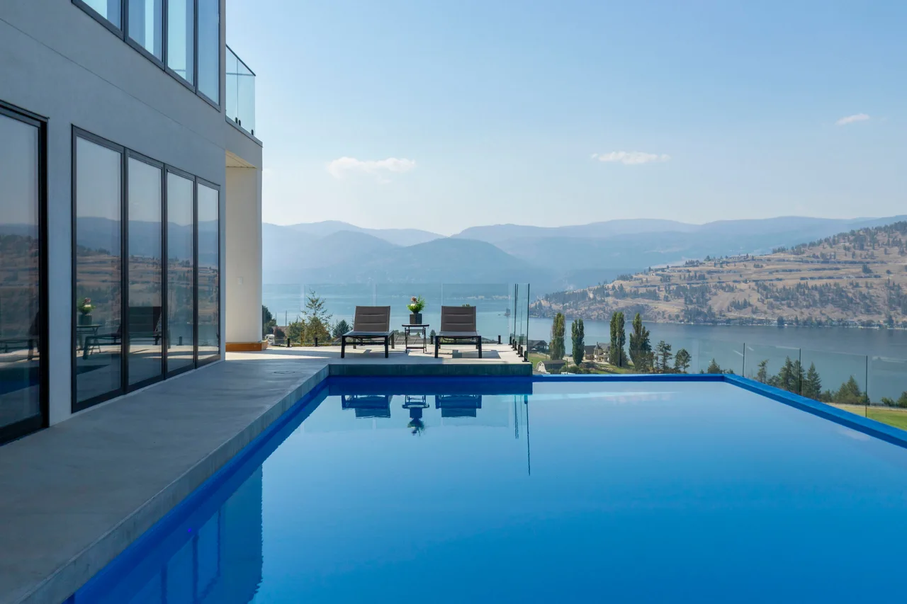 An infinity pool beside a glass-walled home, looking out over Okanagan Lake and the hills